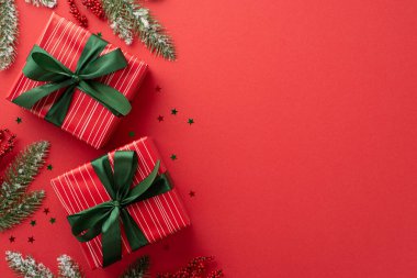 Noel kutlaması kavramı. Yeşil kurdeleli kırmızı hediye kutularının en üst görüntüsü konfeti konfetileri ökseotu üzümleri ve kardaki çam dalları izole edilmiş kırmızı arka planda boş alan.