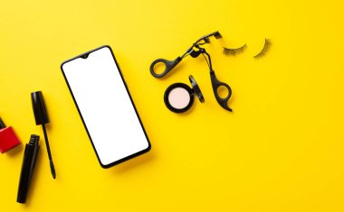Kara Cuma alışveriş konsepti. Akıllı telefon kozmetik maskarası sahte kirpikler göz farı ojeli ve boş alan ile izole edilmiş sarı arka planda kirpik bigudileri.
