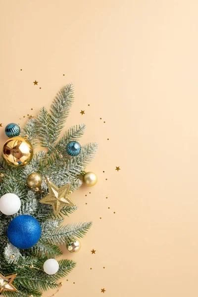 Christmas tree decorations concept. Top view vertical photo of pine branch in frost decorated with white blue gold baubles and confetti on isolated beige background with copyspace