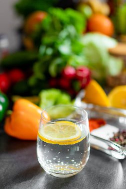 A closeup of a glass with water and lemon on a table full of healthy vegetables