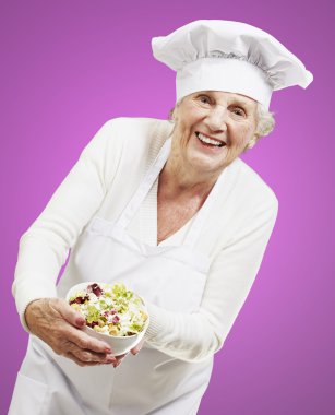 senior woman cook holding a bowl with salad against a pink backg clipart