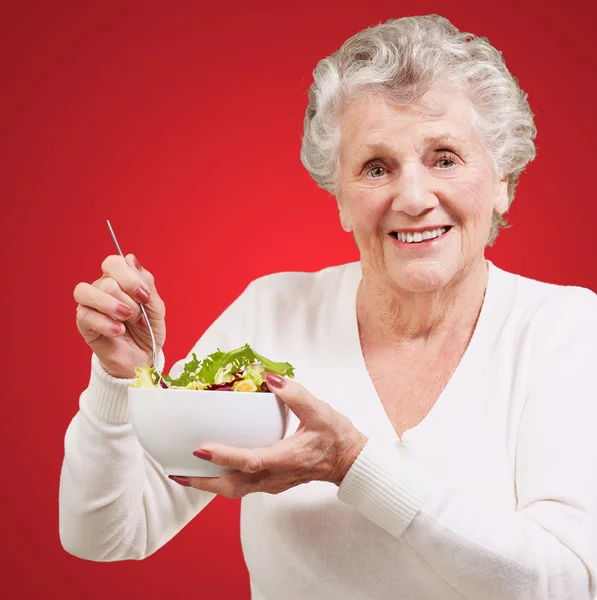 Retrato de mulher idosa comendo salada sobre fundo vermelho — Fotografia de Stock