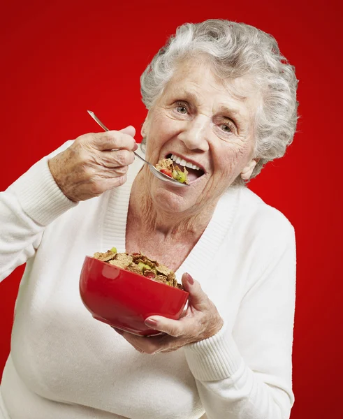 Senior woman eating cereals out of a red bowl against a red back — Stock Photo, Image