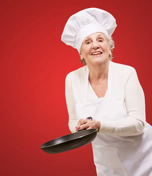 Retrato de una cocinera amigable mujer mayor sosteniendo la sartén sobre ba rojo —  Fotos de Stock