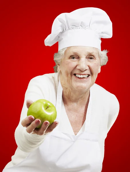 Mujer mayor cocinero ofreciendo una manzana verde contra un fondo rojo —  Fotos de Stock