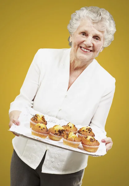 Mujer mayor sonriendo y sosteniendo una bandeja con magdalenas contra una y — Foto de Stock