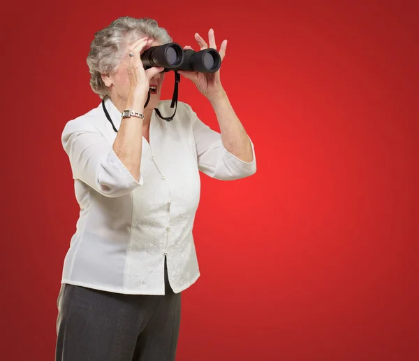 Retrato de mulher sênior olhando através de um binóculos sobre b vermelho — Fotografia de Stock