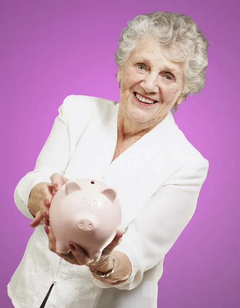 Porträt einer Seniorin, die ein Sparschwein über rosa Hintergrund zeigt — Stockfoto