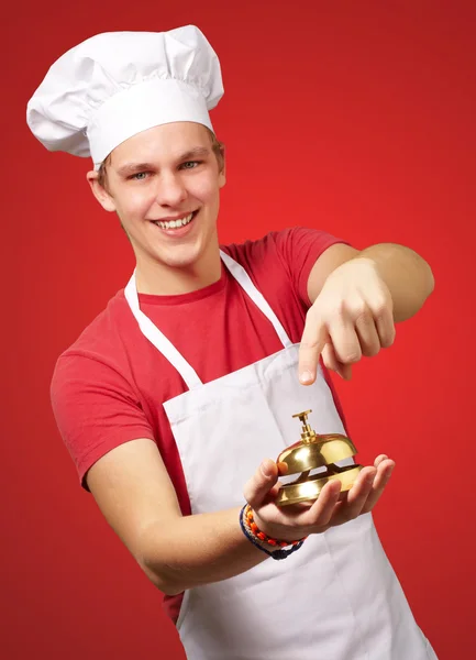 Retrato del joven cocinero presionando una campana dorada sobre el respaldo rojo — Foto de Stock