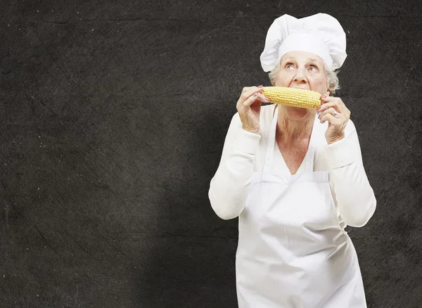 Senior woman cook eating a corncob against a grunge background — Stock Photo, Image