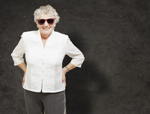 Portrait of senior woman standing wearing sunglasses against a g — Stock Photo, Image