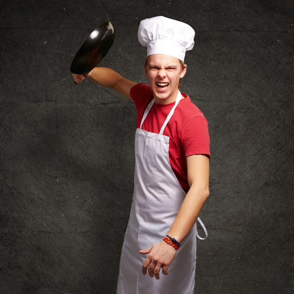 Portret van boze jonge kok man raken met pan tegen een grun — Stockfoto