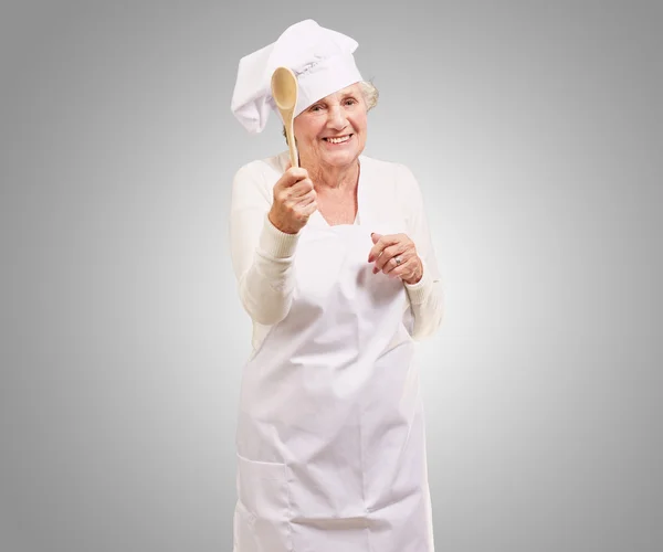 Portrait of senior cook woman holding a wooden spoon over grey b — Stock Photo, Image