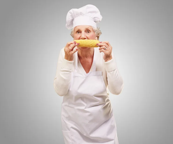 Porträt einer älteren Köchin, die Maiskolben vor grauem Hintergrund isst — Stockfoto