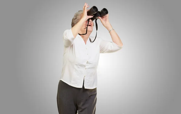 Retrato de mulher idosa olhando através de um binóculos sobre cinza — Fotografia de Stock