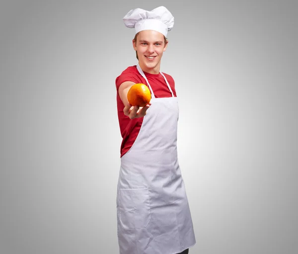 Portrait de jeune cuisinier homme offrant orange sur fond gris — Photo