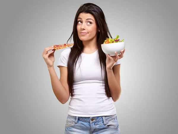 Retrato de mulher jovem escolhendo pizza ou salada contra um b cinza — Fotografia de Stock