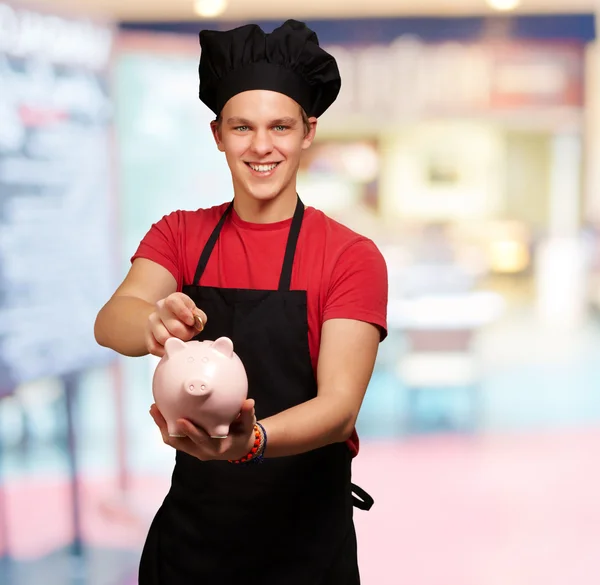 Male chef holding a piggybank and a coin — Stock Photo, Image