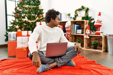 Genç Afro-Amerikalı bir adam dizüstü bilgisayar kullanıyor. Noel ağacının yanında oturuyor. Yan bakmaya çalışıyor, rahat profil pozu veriyor.. 