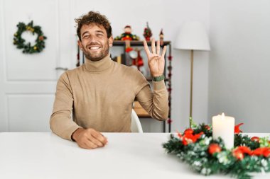 Noel süslemesi masasında oturan sakallı genç yakışıklı dört numarayı gösterip işaret ederken kendinden emin ve mutlu bir şekilde gülümsüyor.. 