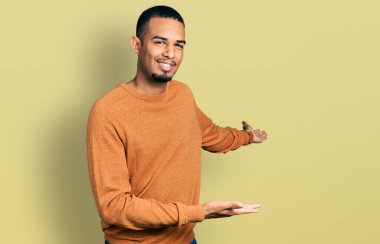 Genç, Afro-Amerikan bir adam, günlük giysiler içinde gülümseyerek içeri girmeye davet ediyor. 