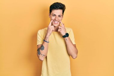Gündelik sarı tişört giyen genç İspanyol adam ağzı açık gülümsüyor, parmakları işaret ediyor ve zorla neşeli bir gülümseme sergiliyor 