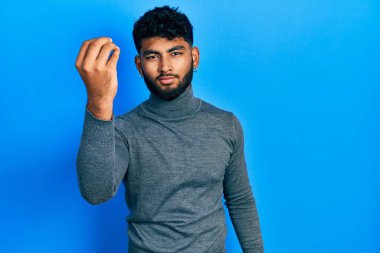 Balıkçı yaka kazak giyen, sakallı Arap bir adam elleriyle ve parmaklarıyla italyan jesti yapıyor. 