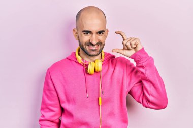 Spor kıyafetleri giyen, kulaklık takan, gülümseyen, kendine güvenen, el hareketi yapan, parmaklarıyla işaret eden ve kamera kullanan genç kel bir adam. ölçüm kavramı. 