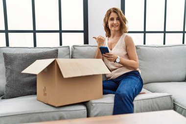 Orta yaşlı bir kadın koltukta oturmuş, teslimat paketini kontrol ediyor. Başparmağını kaldırıp, ağzı açık bir şekilde gülümsüyor. 