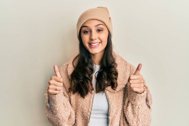 Genç İspanyol kadın yün kazak giyiyor ve kış şapkası takıyor. Elleriyle olumlu jest yapıyor. Gülümsüyor ve mutlu. Neşeli ifade ve kazanan jest. 