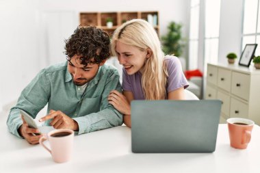 Laptop ve akıllı telefon kullanan genç bir çift evde kahve içiyor..