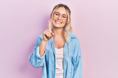 Güzel, genç, sarışın bir kadın, günlük kıyafetler ve gözlük takıyor. Bir numaralı parmağını gösterip işaret ederken, kendinden emin ve mutlu gülümsüyor.. 