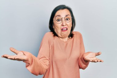 Orta yaşlı İspanyol kadın gündelik kıyafetler giyiyor ve gözlük takıyor. Hiçbir şeyden haberi yok ve elleri ve kolları havada kafası karışmış bir ifade takınıyor. şüphe kavramı. 