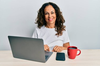 Ofiste çalışan orta yaşlı güzel bir kadın bilgisayarını kullanıyor. Pozitif ve mutlu görünüyor. Kendine güvenen bir gülümsemeyle dişlerini gösteriyor. 