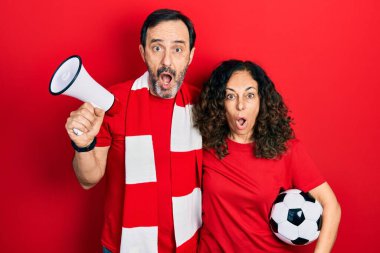 Orta yaşlı bir çift İspanyol kadın ve erkek eşarp takıp tezahürat yapıyorlar ellerinde megafon ve top tutuyorlar ve şaşkınlık ve şaşkınlık ifadesi, korku ve heyecan dolu yüz ifadesiyle şok oluyorlar.. 