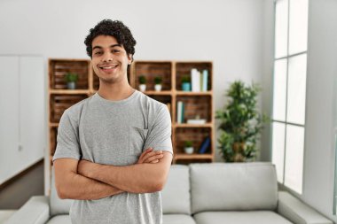 Genç İspanyol adam mutlu gülümsüyor. Kollarını kavuşturup evde duruyor..