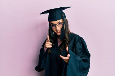 Mezuniyet şapkası ve tören cübbesi takan genç İspanyol kadın mutlu ve komik yüzlü parmaklarını kameraya doğrultuyor. iyi enerji ve titreşimler. 