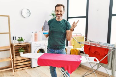 Orta yaşlı, sakallı, evde ütü yapan, gülümseyen, eli ve parmağı kenarda mutlu bir adam. 