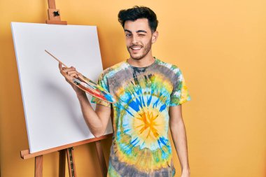 İspanyol asıllı genç adam paletle resim çiziyor, resim sehpası ayakta dikiliyor, kameraya seksi bir ifadeyle bakıyor, neşeli ve mutlu bir yüzle.. 