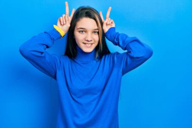 Balıkçı yaka kazak giyen genç esmer kız komik ve çılgın poz veriyor parmakları tavşan kulağı gibi, neşeli gülümsüyor. 