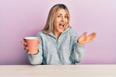 Genç, beyaz bir kadın bir fincan kahve içerek başarıyı mutlu bir gülümsemeyle ve kazanan bir ifadeyle kutluyor. 