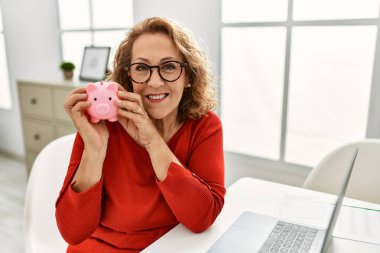 Orta yaşlı beyaz kadın elinde domuz kumbarasıyla evdeki masada oturuyor..