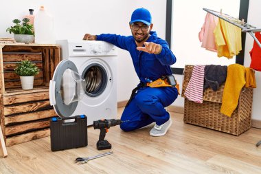 Çamaşır makinesinde çalışan genç Hintli teknisyen seni ve kamerayı parmaklarıyla göstererek mutlu ve pozitif gülümsüyor. 