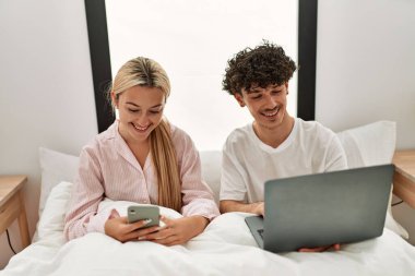 Dizüstü bilgisayar ve akıllı telefon kullanan genç ve güzel bir çift evdeki yatakta oturuyor..