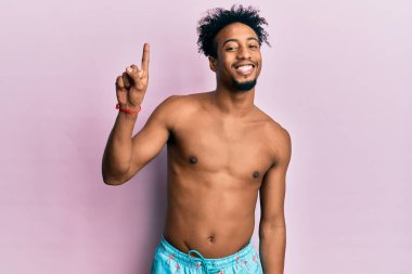 Sakallı, mayo giymiş, gülümseyen, kendinden emin ve mutlu bir şekilde bir numaralı parmağıyla işaret eden Afrikalı genç bir Amerikalı.. 