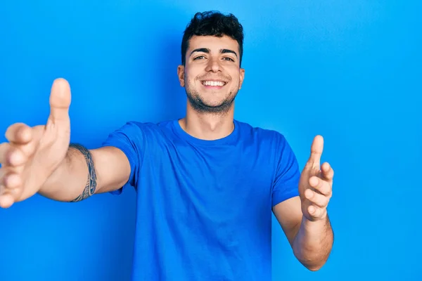カジュアルブルーのTシャツを着た若いヒスパニック系の男が抱擁のために腕を開いて笑顔カメラを見ています 幸せを受け入れる陽気な表情 — ストック写真