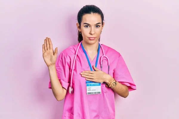Young brunette woman wearing doctor uniform and stethoscope swearing with hand on chest and open palm, making a loyalty promise oath
