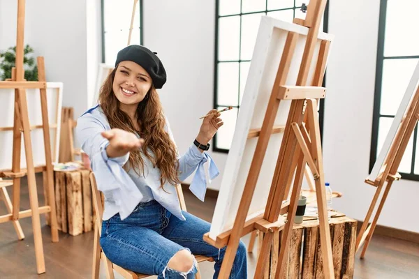 Young hispanic artist woman painting on canvas at art studio smiling friendly offering handshake as greeting and welcoming. successful business.