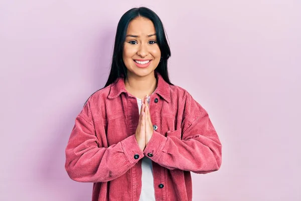 Beautiful hispanic woman with nose piercing wearing casual pink jacket praying with hands together asking for forgiveness smiling confident.