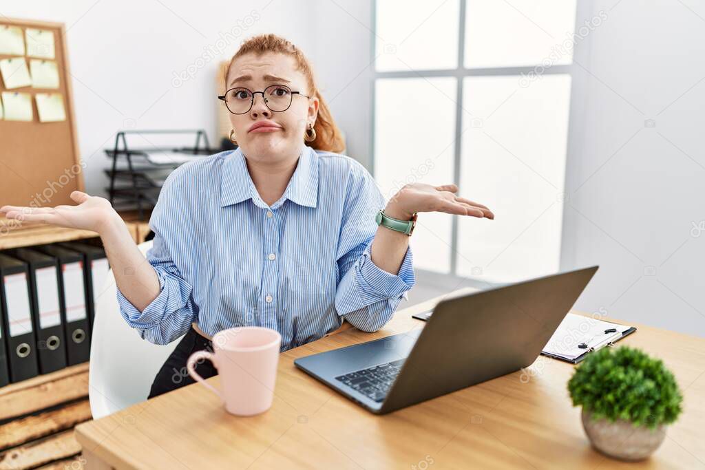 Young redhead woman working at the office using computer laptop clueless and confused expression with arms and hands raised. doubt concept. 